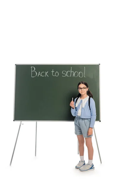Écolière souriante en lunettes tenant de la craie près du tableau noir avec lettrage de retour à l'école sur fond blanc — Photo de stock