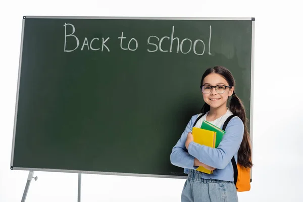 Cheerful schoolkid holding books near chalkboard with back to school lettering isolated on white — Stock Photo