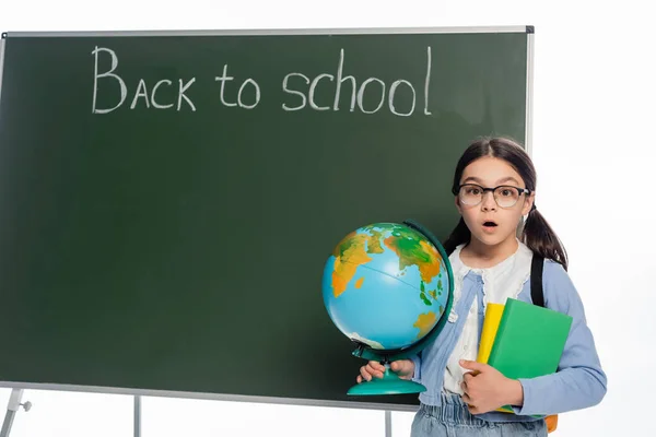 Scioccato scolaro in possesso di libri e globo vicino lavagna con ritorno a scuola lettering isolato su bianco — Foto stock