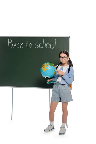 Усміхнений школяр, що вказує на глобус біля крейдяної дошки, спиною до шкільного напису на білому тлі — стокове фото