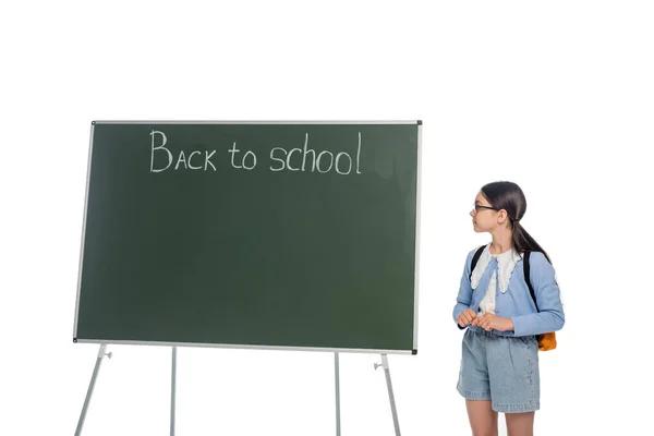 Vue latérale de l'écolier en lunettes regardant le tableau noir avec lettrage de retour à l'école isolé sur blanc — Photo de stock