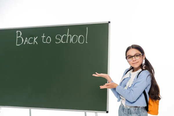Scolaretta in occhiali che punta alla lavagna con scritte di ritorno a scuola isolate su bianco — Foto stock