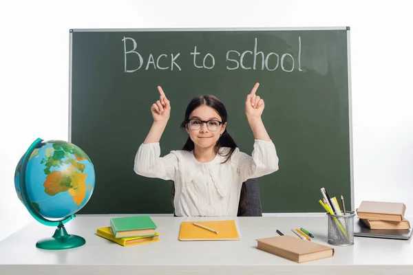 Escolar sonriente apuntando a pizarra con letras de vuelta a la escuela cerca del globo y libros aislados en blanco - foto de stock