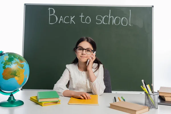 Alumno hablando en smartphone cerca de libros y pizarra con letras de vuelta a la escuela aisladas en blanco - foto de stock