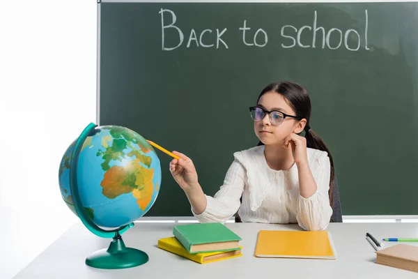 Schulmädchen zeigt auf Globus in der Nähe von Büchern und Kreidetafel mit Schulbuchstaben auf weißem Hintergrund — Stockfoto