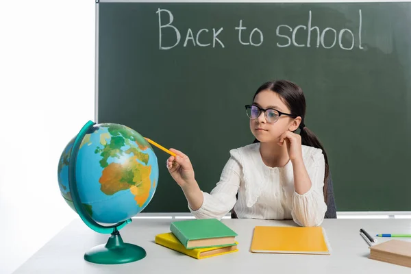 Colegiala apuntando al globo cerca de libros y pizarra con letras de vuelta a la escuela aisladas en blanco - foto de stock