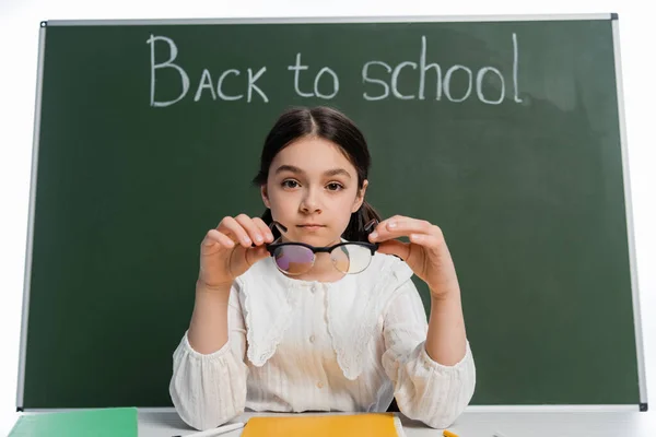 Bambino in età scolare che tiene gli occhiali vicino al taccuino e alla lavagna con iscrizione a scuola isolata su bianco — Foto stock