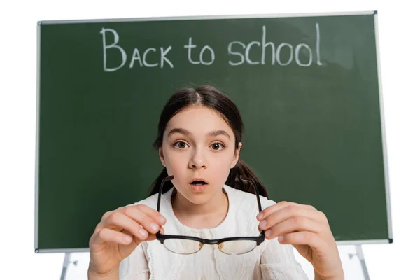 Estudante chocado segurando óculos perto de quadro embaçado com letras de volta para a escola isoladas em branco — Fotografia de Stock
