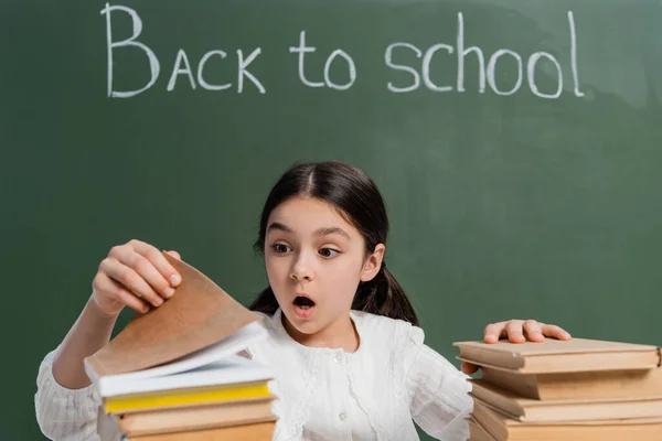 Shocked schoolchild looking at blurred books near back to school lettering on chalkboard — Stock Photo