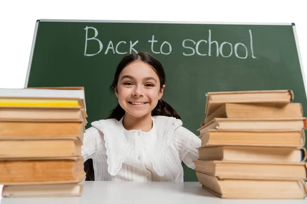 Positives Schulkind blickt in die Kamera neben verschwommenen Büchern und Kreidetafeln mit Schulbuchstaben auf weißem Hintergrund — Stockfoto