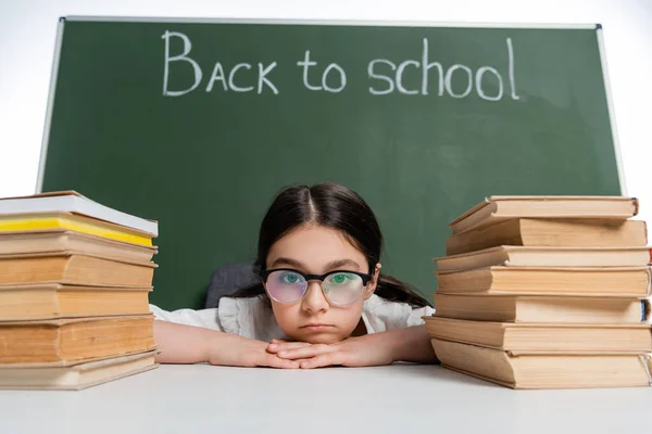 Colegiala molesta en anteojos mirando a la cámara cerca de libros y pizarra con letras de vuelta a la escuela aisladas en blanco - foto de stock