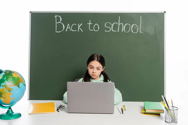 Écolier utilisant un ordinateur portable près des livres et du tableau noir avec lettrage de retour à l'école isolé sur blanc — Photo de stock