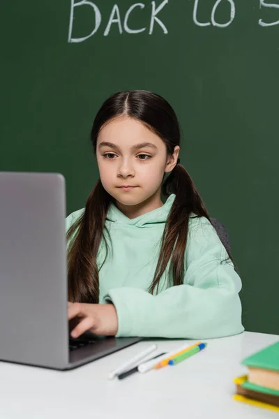 Preteen schoolgirl using laptop near books and chalkboard at background — Stock Photo