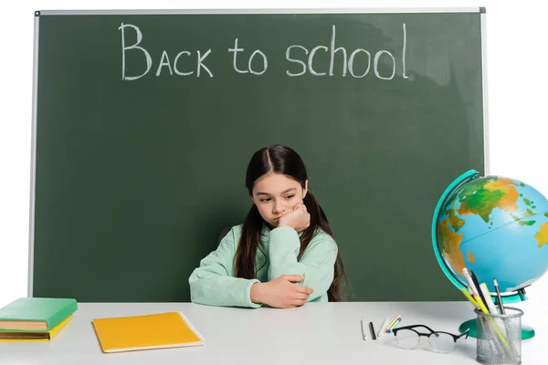 Allievo triste seduto vicino a libri sul tavolo e lavagna con iscrizione a scuola isolato su bianco — Foto stock