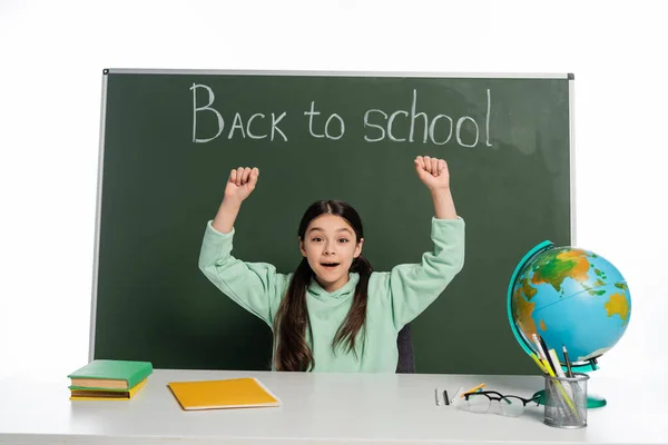 Excited schoolgirl sitting near books and chalkboard with back to school lettering isolated on white — Stock Photo