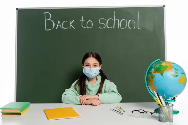 Schoolkid in medical mask sitting near books and chalkboard with back to school lettering isolated on white — Stock Photo