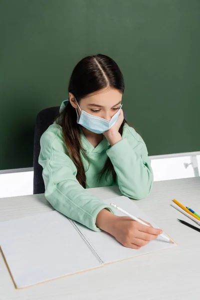 Schoolkid in medical mask writing on notebook near chalkboard isolated on white — Stock Photo