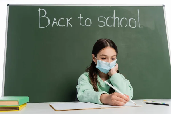 Alumno preadolescente en escritura de máscara médica en un cuaderno cerca de pizarra con letras de regreso a la escuela aisladas en blanco - foto de stock
