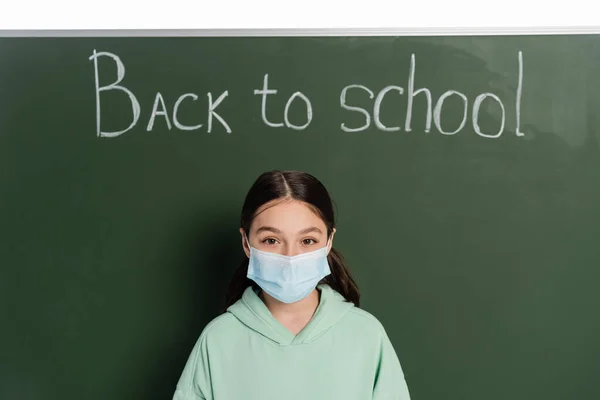 Estudante pré-adolescente em máscara médica olhando para a câmera perto de quadro-negro com volta para a escola lettering isolado no branco — Fotografia de Stock