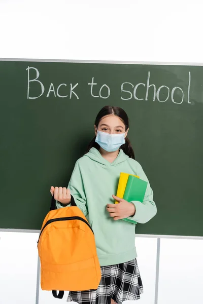 Schoolkid in medical mask holding books and backpack near chalkboard with back to school lettering isolated on white — Stock Photo