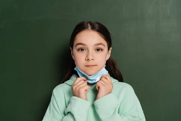 Écolière tenant un masque médical près du tableau noir à l'arrière-plan — Photo de stock