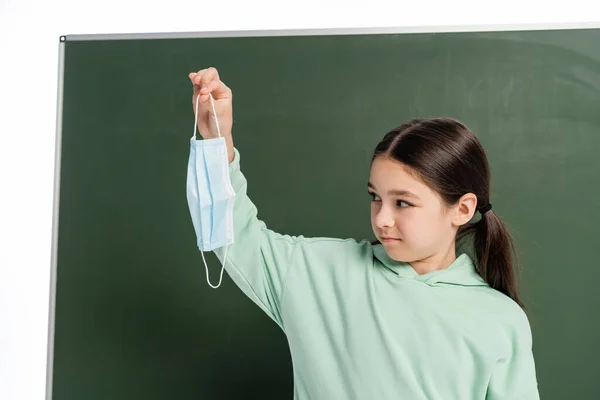 Schulkind blickt auf medizinische Maske in der Nähe von Kreidetafel isoliert auf weiß — Stockfoto