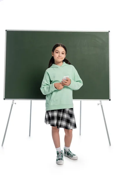 Toute la longueur de l'écolière tenant smartphone près de tableau noir sur fond blanc — Photo de stock