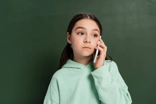 Preteen pupil talking on cellphone near chalkboard at background — Stock Photo