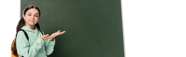 Smiling schoolkid with backpack pointing at chalkboard isolated on white, banner — Stock Photo
