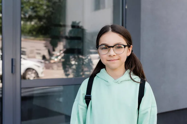 Усміхнена школярка в окулярах дивиться на камеру біля будівлі на відкритому повітрі — стокове фото