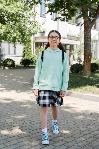 Enfant d'âge préscolaire avec sac à dos et lunettes regardant la caméra dans la rue urbaine — Photo de stock