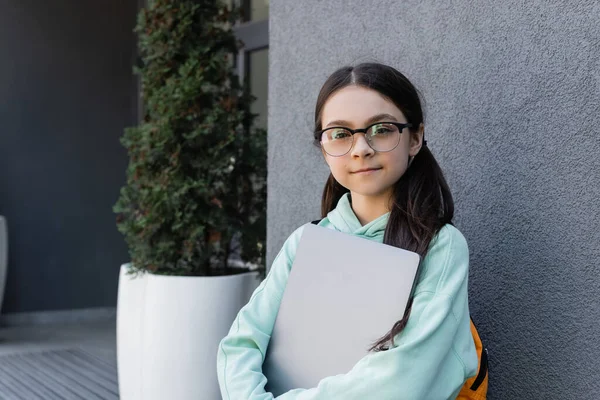 Portrait of schoolchild in eyeglasses holding laptop on urban street — Stock Photo
