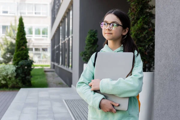 Preteen schoolchild in eyeglasses holding laptop on urban street — Stock Photo