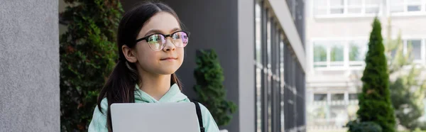 Preadolescente escolar en gafas con portátil y mirando hacia otro lado en la calle urbana, pancarta - foto de stock