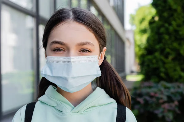 Retrato de colegiala en máscara médica mirando la cámara en la calle urbana - foto de stock