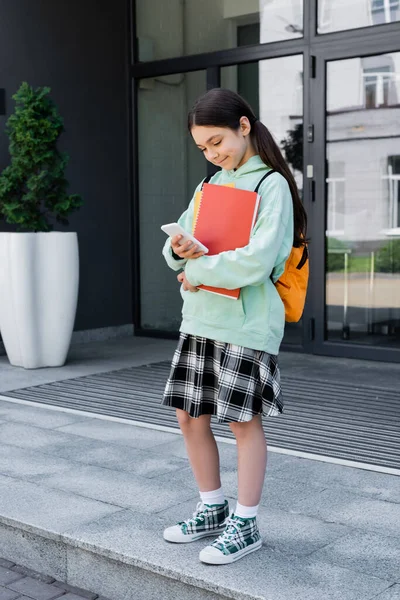 Улыбающийся школьник с ноутбуками, использующий мобильный телефон возле здания на открытом воздухе — стоковое фото