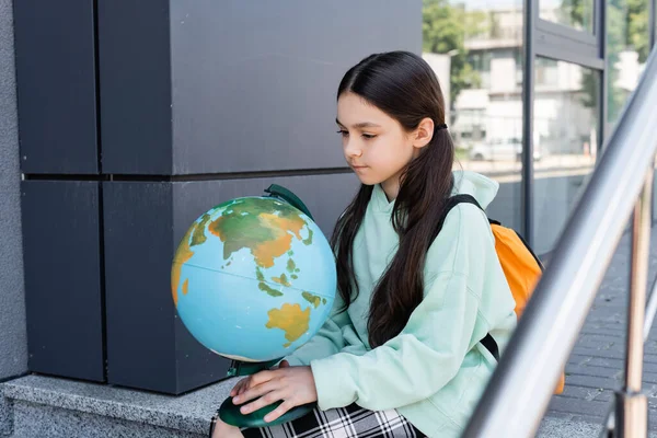 Preteen schoolgirl with backpack looking at globe near building on urban street — Stock Photo