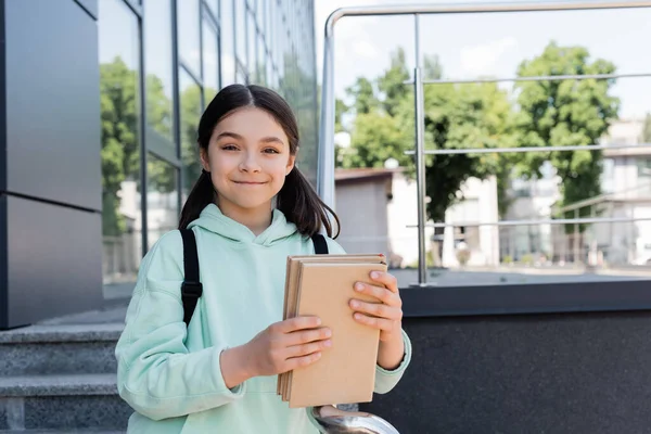 Écolier souriant avec sac à dos tenant des livres près du bâtiment à l'extérieur — Photo de stock