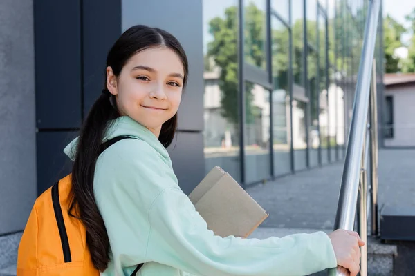 Positive Schülerin mit Rucksack und Büchern, die in der Nähe verschwommener Bücher in die Kamera schauen — Stockfoto