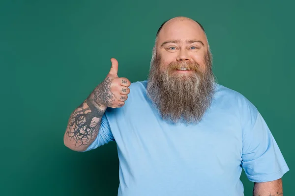 Hombre barbudo alegre con tatuajes mostrando el pulgar hacia arriba aislado en verde - foto de stock