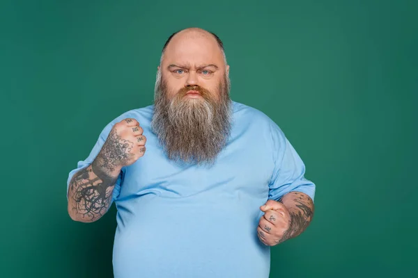 Angry overweight man with tattoos and beard showing clenched fists isolated on green — Stock Photo