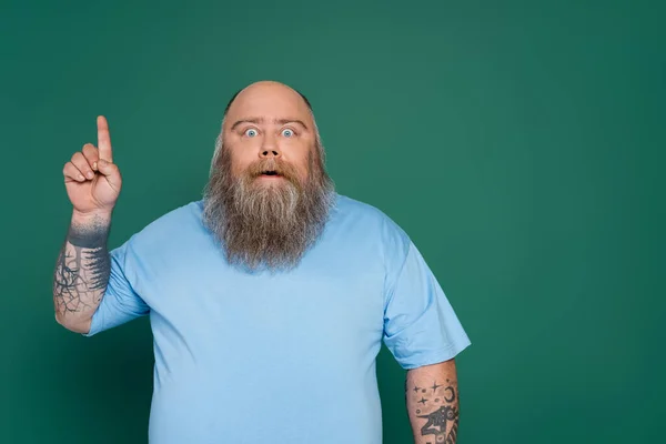 Surpreso homem com excesso de peso com barba apontando para cima com o dedo isolado no verde — Fotografia de Stock