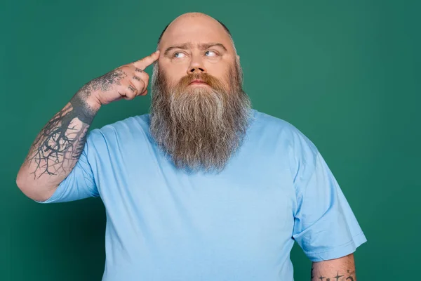 Tatuado más tamaño hombre tocando la cabeza y mirando hacia otro lado mientras piensa aislado en verde - foto de stock
