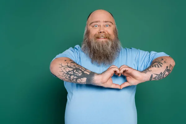 Happy plus size man with beard and tattoos showing heart sign isolated on green — Photo de stock