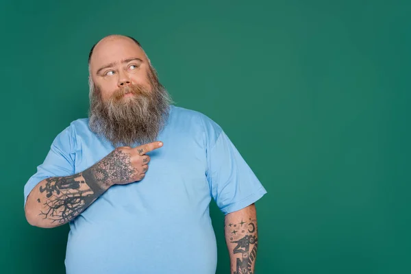Homme en surpoids réfléchi avec des tatouages pointant du doigt et regardant loin isolé sur vert — Photo de stock
