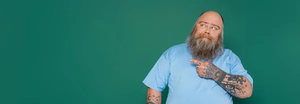 Homem com excesso de peso pensativo com barba olhando para longe e apontando com o dedo isolado no verde, banner — Fotografia de Stock