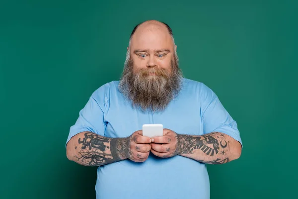 Emocionado más hombre de tamaño con barba charlando en el teléfono inteligente aislado en verde - foto de stock