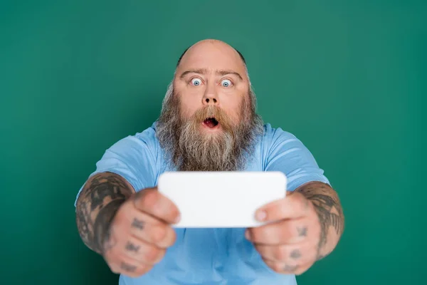 Astonished overweight man with beard and tattoos taking selfie on blurred smartphone isolated on green — Stock Photo