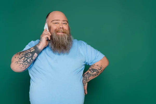 Щасливий і бородатий плюс розмір чоловік стоїть з рукою на талії і розмовляє на мобільному телефоні ізольовано на зеленому — стокове фото