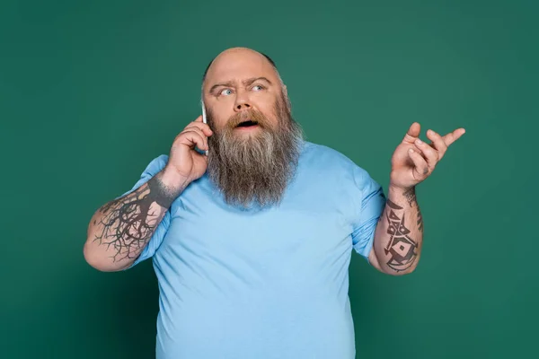 Hombre con sobrepeso reflexivo tatuajes hablando en el teléfono inteligente y señalando con el dedo aislado en verde - foto de stock